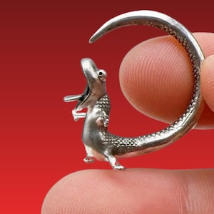 Puede incluir: Un anillo plateado con un diseño detallado de un caimán. El caimán está posicionado junto a una banda curva, con la boca abierta. El anillo se sostiene sobre un fondo rojo.
