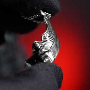 Puede incluir: Un collar con colgante plateado con un diseño de rostro estilizado. El colgante cuelga de una cadena delicada. La cara tiene detalles y patrones geométricos. El fondo es un degradado de rojo.