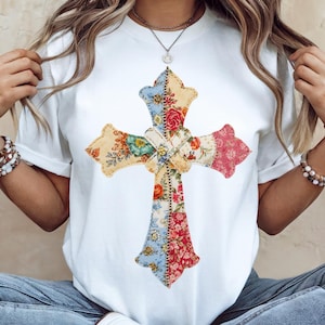 以下が含まれることがあります： パッチワークの十字架のデザインが施された白いTシャツ。十字架は、青、赤、黄、緑の色合いのフローラル生地で作られています。十字架にはステッチの輪郭と中央にリボンが付いています。このシャツはカジュアルなスタイルです。