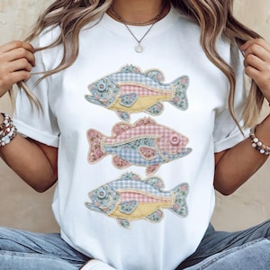 May include: White t-shirt with three decorative fish appliqués. Each fish has a unique patchwork design with floral, gingham, and lace details in pastel colours. The fish are arranged vertically on the shirt.