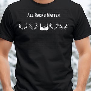 May include: Black t-shirt with the text "ALL RACKS MATTER" in white. The shirt features white illustrations of antlers and a bra. The background is a blurred winter scene.