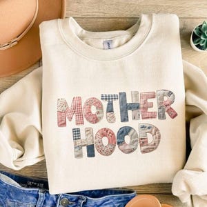 May include: Cream-colored sweatshirt with the word "MOTHERHOOD" in patchwork letters. The letters are made of various patterned fabrics in shades of red, blue, and white. A tan hat and blue jeans are also visible.