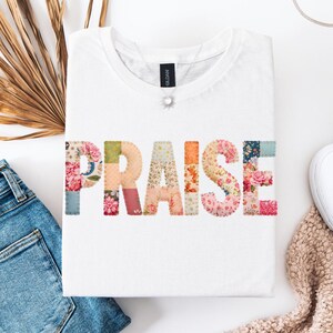 Puede incluir: Camiseta blanca con la palabra "PRAISE" en letras de patchwork floral colorido. La camiseta se muestra con unos vaqueros azules, una hoja de palma seca y zapatos blancos. Se ve un collar plateado.