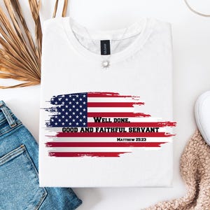 May include: White t-shirt with a graphic of the American flag and the text "Well Done, Good and Faithful Servant Matthew 25:23". The shirt is folded and placed on a white surface next to a pair of blue jeans and white shoes.