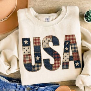 May include: Cream-colored sweatshirt with the word "USA" appliqued on the front. The letters are made of patchwork fabric in red, white, and blue patterns, including stars and plaid. A tan hat and denim shorts are visible.