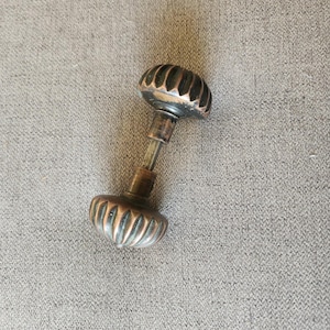 May include: A pair of antique-style door knobs featuring a fluted, pumpkin-shaped design with a dark, weathered copper finish. The two knobs are connected by a central metal spindle, showcasing a metallic texture with visible patina and dark accents.