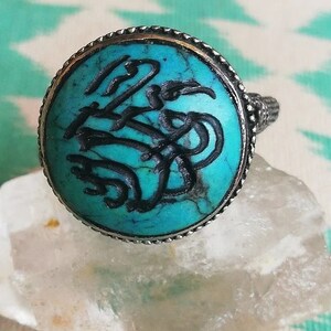 Op de afbeelding: Een zilveren ring met een turquoise steen in een ronde zetting. De steen is versierd met ingewikkelde zwarte gravures.