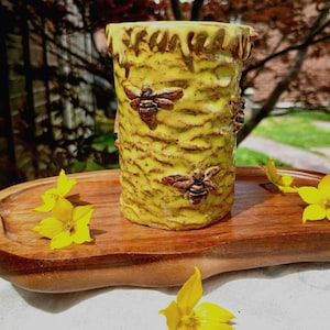May include: A cylindrical yellow ceramic vessel with a textured honeycomb surface and raised bee figures, featuring a dripping glaze effect at the top rim. The vessel rests on a smooth, dark brown wooden tray adorned with several small yellow flowers.