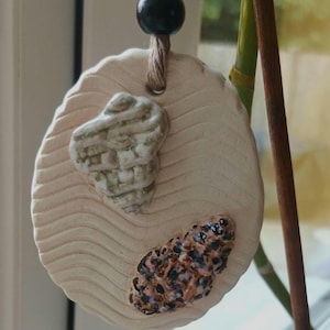 May include: An oval-shaped ceramic ornament with a wavy, textured surface in a light beige tone. It features two attached shell-like decorations: one pale green and one brown with black spots. A jute twine loop with a single black bead hangs from the top.