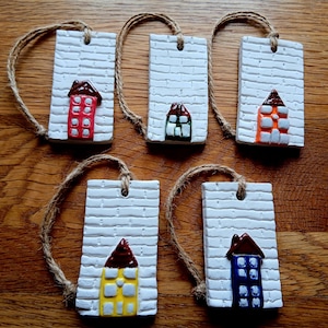 May include: Five rectangular white clay ornaments with a textured brick pattern, each featuring a small house with a brown roof. The houses are painted in red, green, orange, yellow, and blue. Each ornament includes a brown jute twine hanging loop.