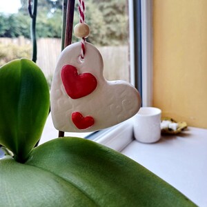 May include: A heart-shaped ornament made of off-white clay with a textured surface featuring embossed heart patterns. Two raised, glossy red hearts are attached to the front. It hangs from a red and white baker's twine string with a wooden bead.