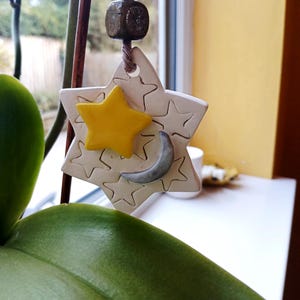 May include: A ceramic star-shaped ornament hanging from a brown twine string with a dark bead. The white star features embossed star outlines, a glossy yellow star accent, and a smooth grey crescent moon shape attached to the front surface.