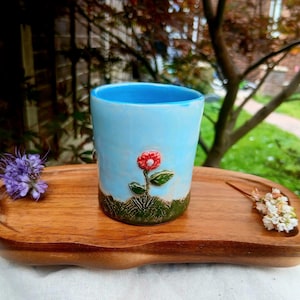 May include: A light blue ceramic cup with a textured green base and a small red flower appliqué, resting on a smooth, oblong wooden tray. The cup features a glossy finish, while the tray displays a natural wood grain and curved edges.