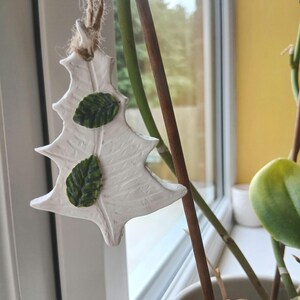 May include: A white, tree-shaped hanging ornament made of a clay-like material with a textured surface. Two small, dark green, detailed leaves are attached to the front. The ornament hangs from a piece of natural brown twine tied at the top.
