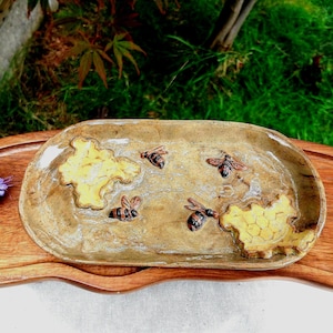 May include: A rectangular ceramic tray with a speckled tan glaze featuring three raised bee figures and two yellow honeycomb patterns. The tray rests on a dark brown wooden serving board with a wavy edge, accompanied by small purple and white flowers.