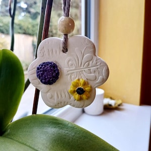 May include: A white, flower-shaped ceramic ornament hanging from a brown twine string with a speckled beige bead. The ornament features embossed leaf patterns, a small purple textured circular accent, and a yellow flower with a black centre.