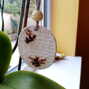 May include: A circular white ceramic ornament featuring a textured honeycomb pattern and two small, detailed bees with black and yellow markings. The ornament hangs from a brown twine cord accented with a single, smooth, light yellow wooden bead.