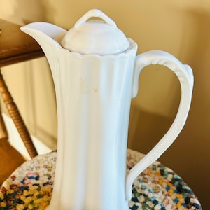 Puede incluir: Cafetera de porcelana blanca con tapa y asa. La cafetera tiene un diseño acanalado y una base redonda.