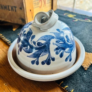 May include: A ceramic butter dish with a domed lid and a matching base. The lid is decorated with blue floral patterns on a white background. The dish has a small, looped handle on top. The base is a light brown color.