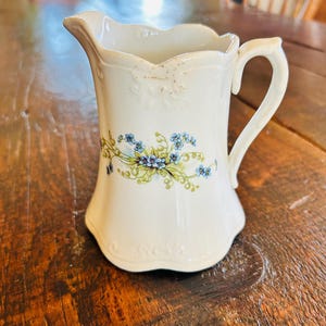 May include: A white porcelain creamer with a scalloped rim and handle. It features a floral design with blue forget-me-not flowers and green leaves.