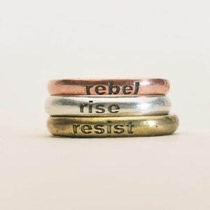 May include: Three stacked metal rings with the words "rebel", "rise", and "resist" stamped on them. The top ring is copper, the middle ring is silver, and the bottom ring is gold. The rings are on a light background.