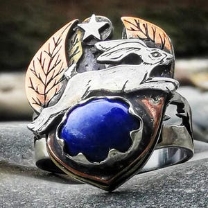 May include: A silver ring with a leaping rabbit design and a blue gemstone. The ring has copper and gold accents, a star, and leaf details. The ring's design is intricate and detailed.