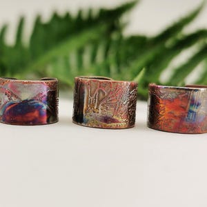 May include: Three handcrafted rings with a textured, iridescent finish. The rings display a blend of colours, including purple, blue, and orange, with a rustic, metallic appearance. The rings are set against a white surface with green foliage in the background.