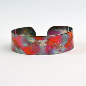 Puede incluir: Una pulsera de cobre con un acabado colorido e iridiscente. La pulsera está decorada con un patrón repetido de siluetas de libélulas.