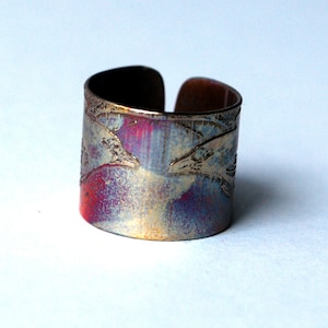 Op de afbeelding: Een brede koperen ring met een getextureerd oppervlak en een blauwe, rode en gouden patina. De ring is gemaakt van metaal en heeft een uniek ontwerp.