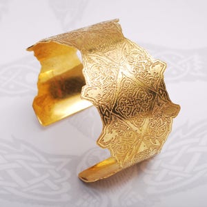 May include: A gold-toned metal cuff bracelet with an intricate, embossed design. The design features a repeating pattern of stylized shapes and swirls.
