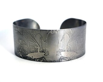 Puede incluir: Una pulsera ancha de puño en tono plateado con un diseño grabado de un pájaro en vuelo. La pulsera tiene un acabado de metal cepillado y una forma ligeramente curvada, con el diseño del pájaro envolviendo la muñeca.