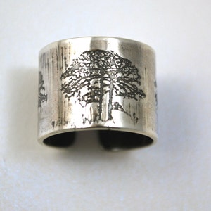 Puede incluir: Un brazalete ancho plateado con un acabado texturizado. El brazalete presenta un diseño grabado de un árbol, con ramas y follaje detallados. El brazalete está abierto en la parte inferior, lo que permite un ajuste ajustable.