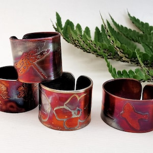 May include: Four copper napkin rings with nature-inspired designs. One ring features a dragonfly, another a tree, a third a flower, and the last a bird. The rings have a reddish-brown and purple patina.