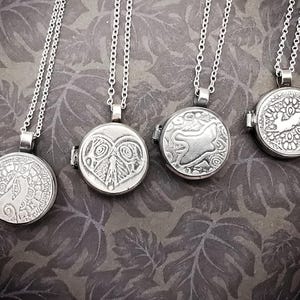 May include: Four silver locket necklaces with detailed designs. Each circular pendant displays a different embossed image: a bird, a heart with leaves, a frog, and a rabbit. The lockets are suspended from fine chain necklaces.