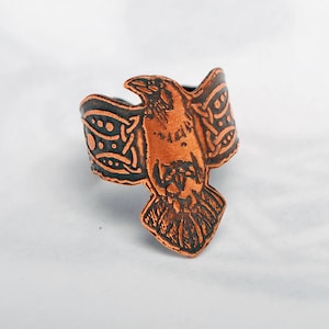 May include: A copper ring with a detailed design of a raven with its wings spread. The ring is a dark copper color with a black patina.