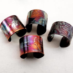 May include: Four handcrafted metal cuff bracelets with unique designs and iridescent colorations. Each cuff features a different etched pattern, including a tree, floral, and abstract designs. The cuffs are open-ended, allowing for easy wear.