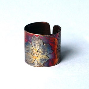 May include: A copper-colored adjustable ring with a floral design. The ring features a vibrant color palette of red, blue, and gold hues. The floral pattern is etched onto the surface, adding texture and detail to the jewelry.