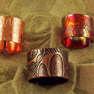 May include: Three copper rings with different embossed patterns. The rings are on a textured brown surface.