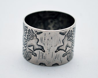 Könnte beinhalten: Ein silberner Ring mit zwei Vögeln im Flug, die sich gegenüberstehen. Der Ring hat eine gebürstete Metallstruktur und ein breites Banddesign. Die Vögel sind aufwendig detailliert. Der Ring ist ein breites Band.