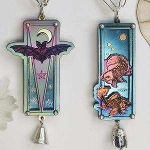 May include: Two iridescent pendants with silver chains. One features a bat, moon, and stars, with a bell charm. The other has two goldfish and bubbles, with a charm. Both have a rectangular frame.
