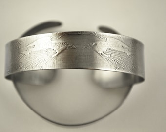 Puede incluir: Una pulsera de metal plateado con un diseño grabado detallado de dos pájaros que se miran.