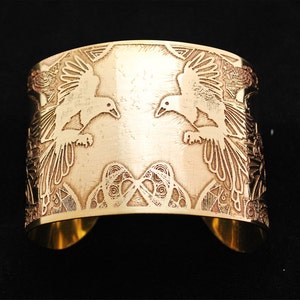 May include: A gold-toned metal cuff bracelet with an intricate design featuring two birds in flight. The birds are facing each other and have detailed wings and bodies. The background of the bracelet is filled with a geometric pattern.