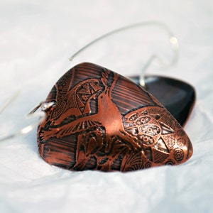 Puede incluir: Pendientes de color cobre con intrincados diseños grabados. Los pendientes tienen una forma única y presentan un patrón detallado de figuras y formas geométricas. Los pendientes están suspendidos de alambres plateados.