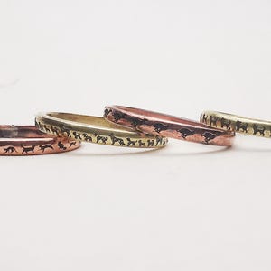 Op de afbeelding: Vier metalen ringen in koper, goud en roségoud tinten. Elke ring heeft een herhalend silhouetontwerp van rennende honden. De ringen worden gepresenteerd op een wit oppervlak.