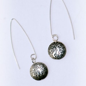 May include: A pair of silver dangle earrings with a circular design. Each earring features a round, silver charm with a detailed, etched pattern.