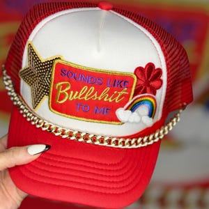 May include: Red and white trucker hat with a gold chain, a star, a rainbow, and a patch that says "Sounds Like Bullshit To Me".