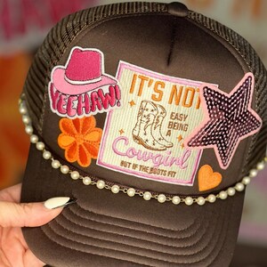 May include: Brown trucker hat with a pearl trim and various patches, including a pink star, a pink heart, a flower, a pink cowboy boot patch that says "It's not easy being a cowgirl, but if the boots fit", and a pink hat with the word "Yeehaw!"