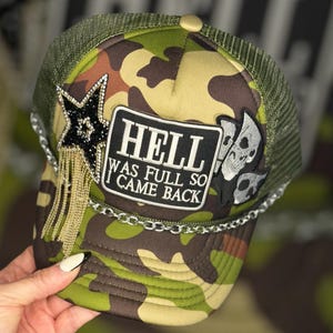 May include: A green camouflage trucker hat with a black star and a patch that reads "HELL WAS FULL SO I CAME BACK". The hat also has a silver chain and a skull patch.