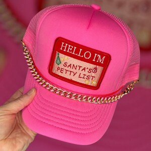 May include: Bright pink trucker hat with a mesh back and a curved brim. The hat features a rectangular patch that reads "HELLO I'M SANTA'S PETTY LIST" in white text on a red background. A gold chain is attached to the hat.