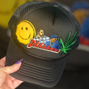 May include: Black trucker hat with a yellow smiley face, a green marijuana leaf, and an embroidered patch that says "Daaamn!" with two cartoon figures.